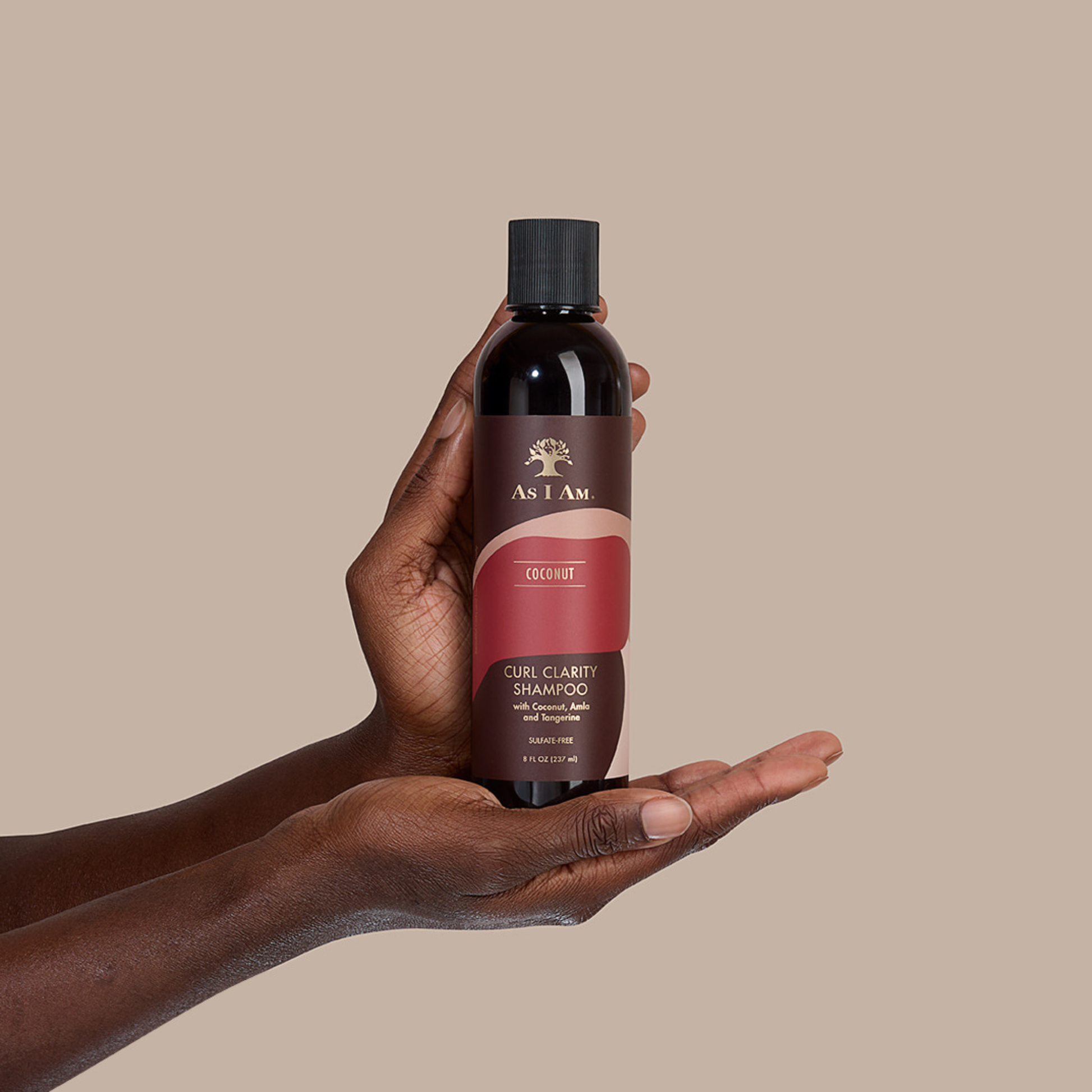 Hand met een fles As I Am Coconut Curl Clarity Shampoo tegen een beige achtergrond