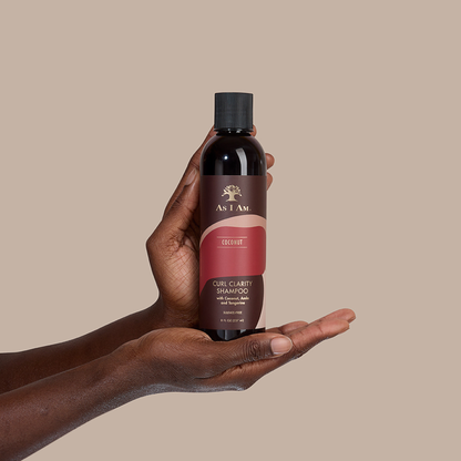 Hand met een fles As I Am Coconut Curl Clarity Shampoo tegen een beige achtergrond